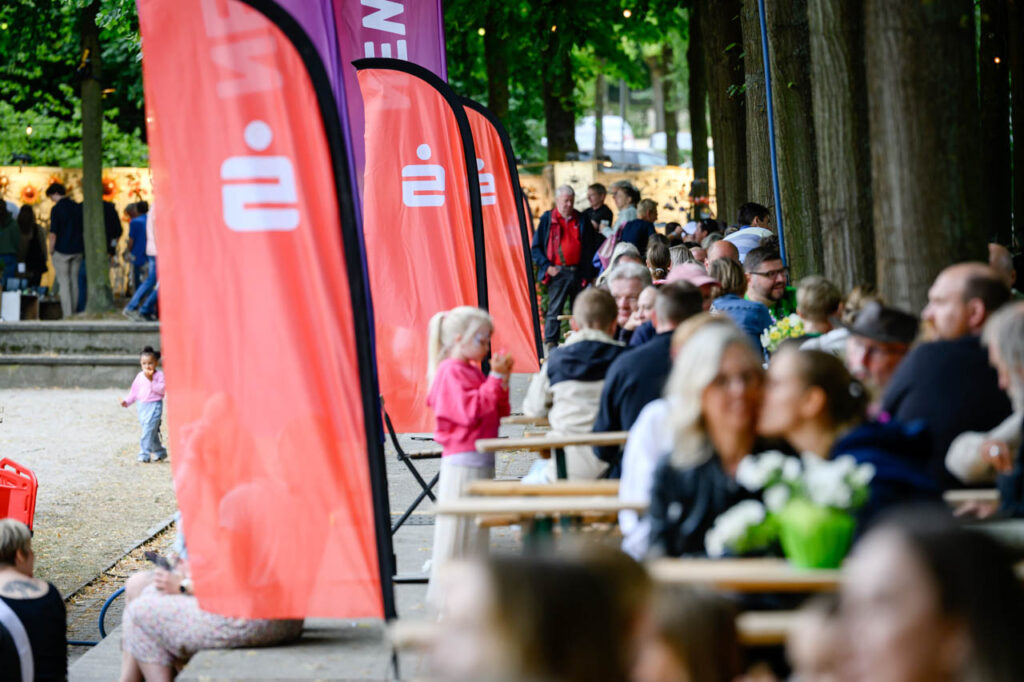 Viele Gäste sitzen an langen Tischen unter Bäumen im Gastronomiebereich beim Fest am See.