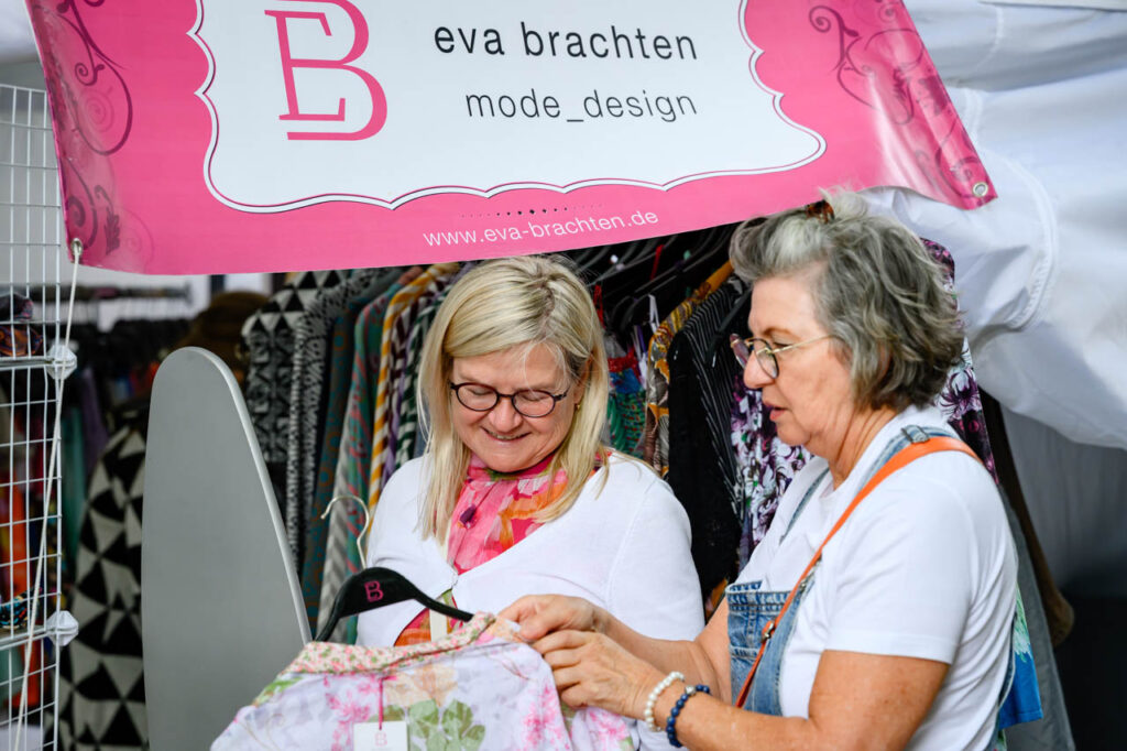 Zwei Besucherinnen schauen sich Kleidung am Stand von Eva Brachten auf dem Heimatliebe Markt beim Fest am See an.