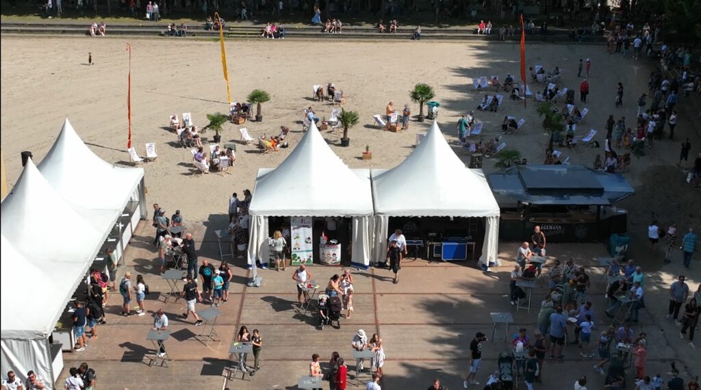 Von oben ist das Festgelände im Schlosspark Wickrath zu sehen. Menschen sitzen in Liegestühlen im Sandbereich, während sich rund um die weißen Zelte weitere Gäste aufhalten und bewegen.