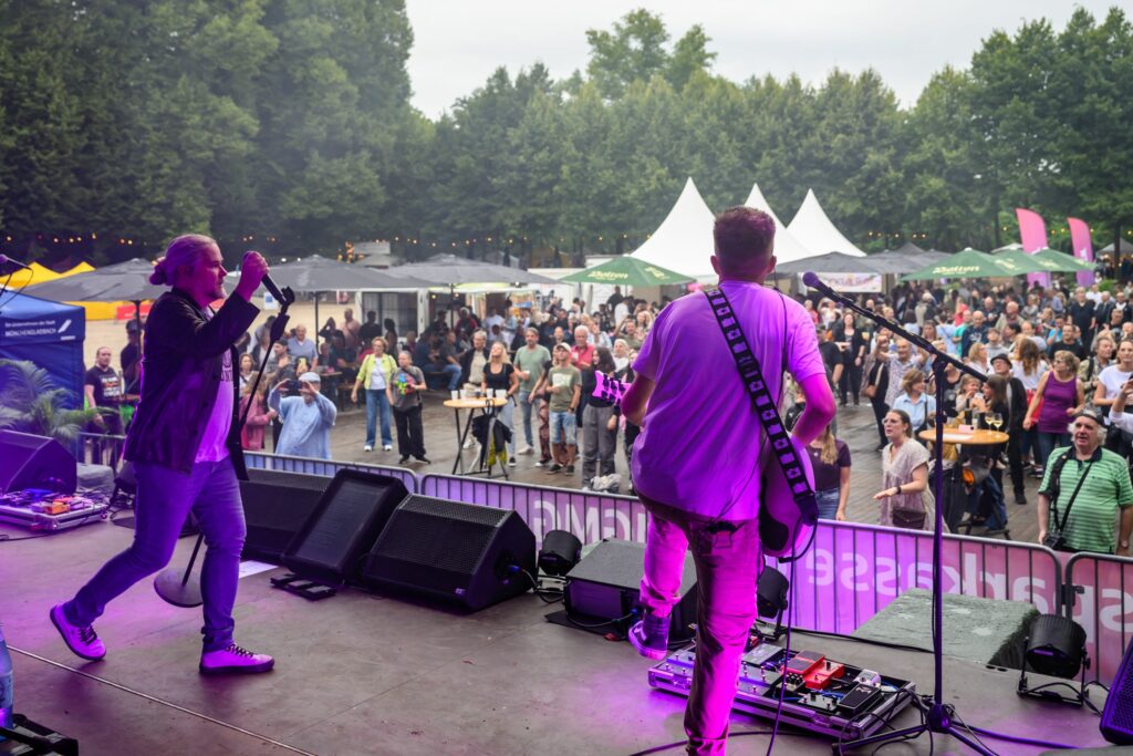 Blick von der Bühne auf das Publikum. Zwei Musiker stehen vorne, Menschen verteilen sich vor der Bühne und zwischen Ständen.