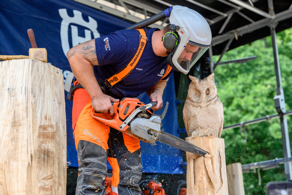 Ein Mann mit Schutzkleidung und Helm arbeitet mit einer Motorsäge an einer Holzskulptur in Form einer Eule. Holzspäne fliegen durch die Luft, während Zuschauer im Hintergrund stehen.