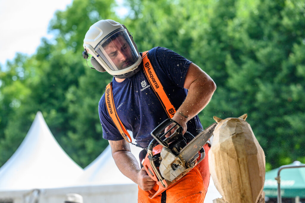 Ein Mann mit Schutzhelm und Motorsäge bearbeitet eine Holzfigur auf einer Bühne. Im Hintergrund stehen Zelte und Zuschauer, die die Vorführung beobachten.