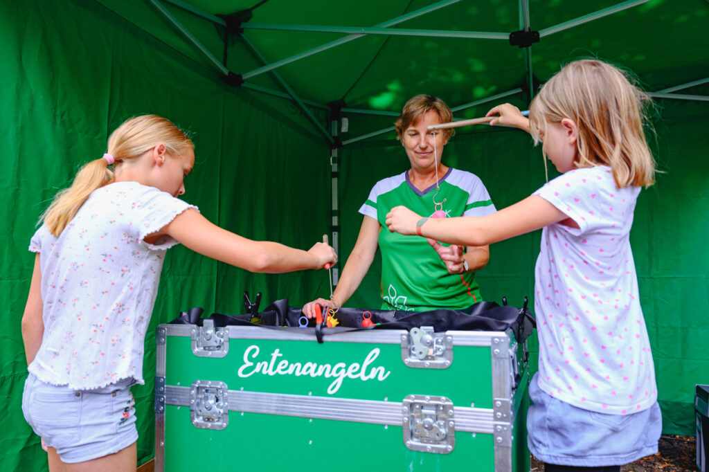 Zwei Kinder angeln mit kleinen Stäben nach Spielzeugenten aus einer Kiste an einem Stand. Eine Frau betreut das Spiel und schaut ihnen zu.