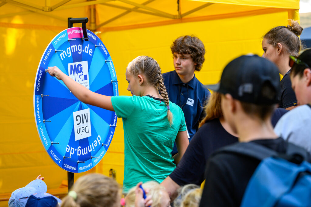 Ein Mädchen dreht an einem bunten Glücksrad mit der Aufschrift mgmg.de, während andere Kinder und Jugendliche zuschauen. Die Szene spielt sich unter einem gelben Pavillon ab.