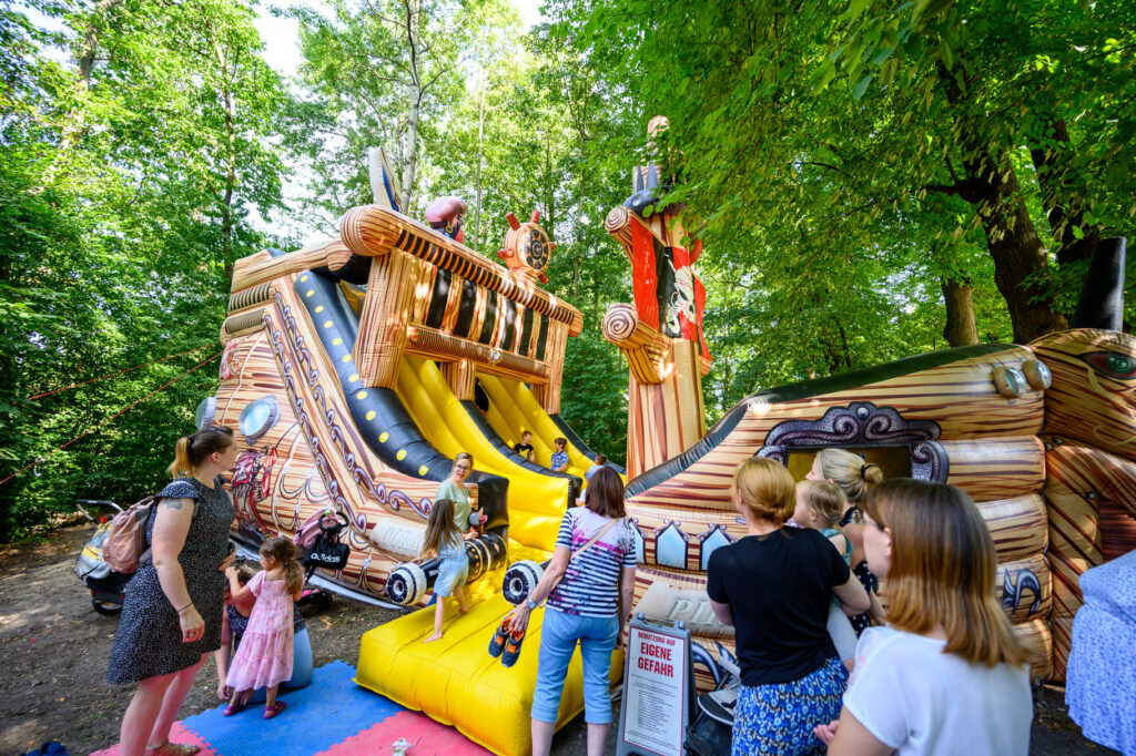 Kinder spielen auf einer großen aufblasbaren Piratenschiff-Rutsche im Wald. Eltern stehen daneben und schauen zu, während die Kinder die Rutsche nutzen.