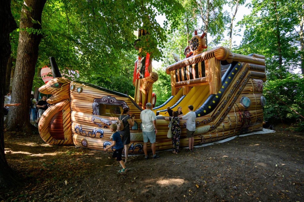 Eine große Piratenschiff-Hüpfburg steht im Grünen. Kinder spielen darauf, während Erwachsene am Rand stehen und das Geschehen beobachten.
