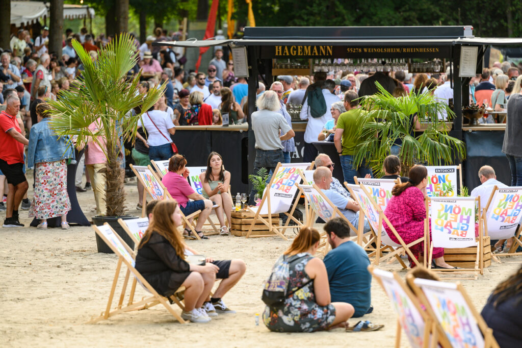 Menschen sitzen entspannt auf Liegestühlen im Sand vor einer Bar. Im Hintergrund stehen viele Besucherinnen und Besucher an einem Getränkestand.