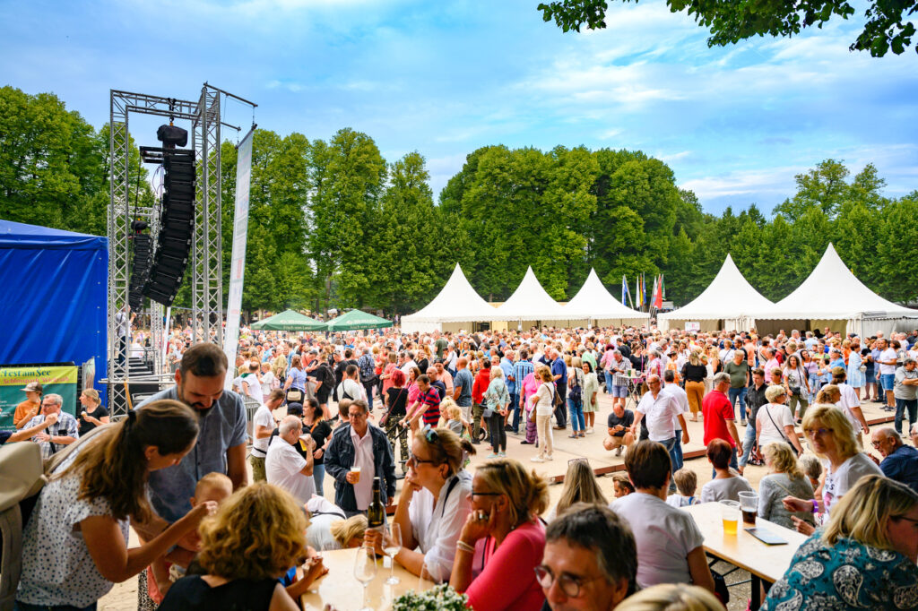 Eine große Menschenmenge steht vor einer Bühne im Park. Im Vordergrund sitzen und stehen Besucherinnen und Besucher an Tischen mit Getränken.
