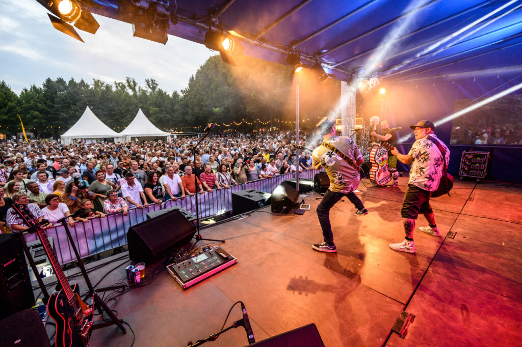 Eine Band tritt auf einer Bühne vor einem großen Publikum auf. Die Musiker spielen Gitarre und stehen unter buntem Bühnenlicht.