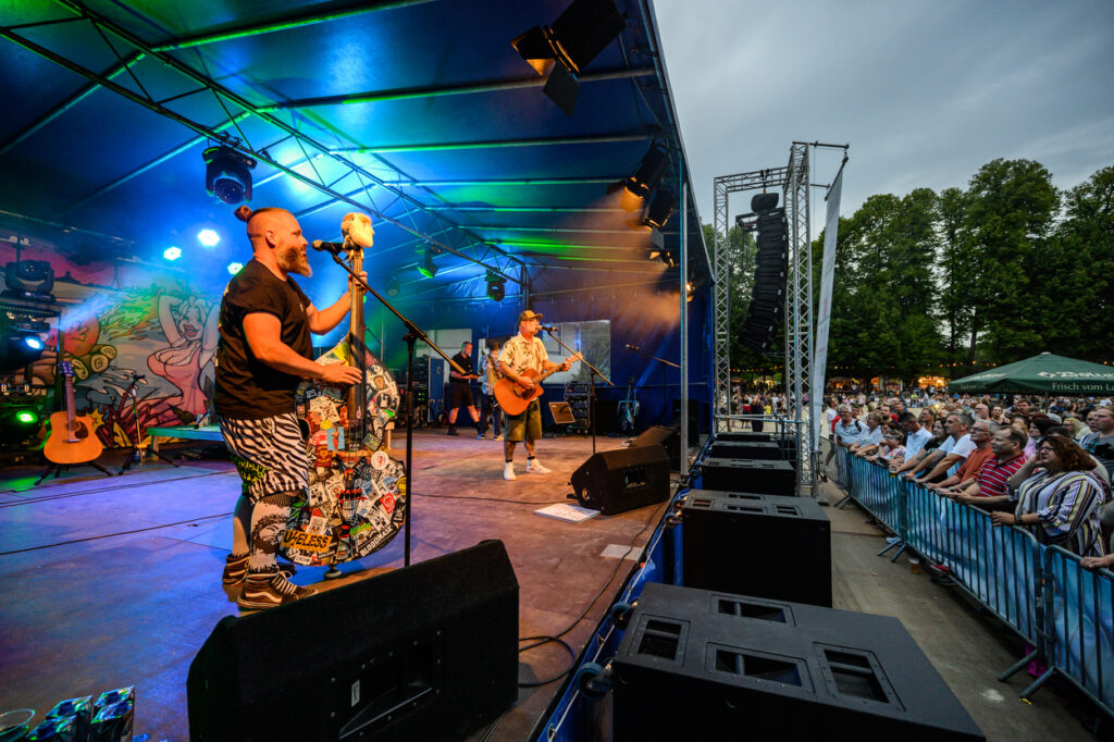 Eine weitere Perspektive auf die Bühne zeigt Musiker während eines Live-Auftritts. Das Publikum steht dicht gedrängt vor der Bühne und schaut zu.