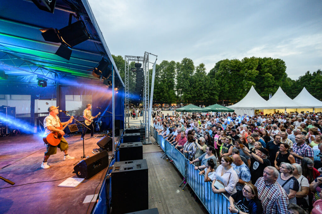 Eine Band spielt auf einer Bühne im Schlosspark Wickrath vor einer großen Menschenmenge. Viele Besucherinnen und Besucher stehen dicht gedrängt vor der Absperrung und verfolgen das Konzert. Im Hintergrund sind Zelte und Bäume zu sehen, die die Atmosphäre des Fest am See in Mönchengladbach prägen.