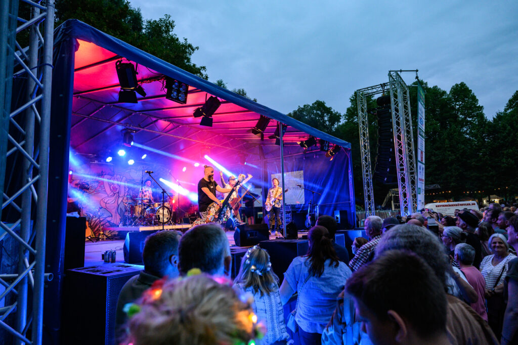 Eine Band spielt auf einer beleuchteten Bühne im Schlosspark Wickrath, während sich davor viele Besucherinnen und Besucher versammeln. Bunte Scheinwerfer tauchen die Bühne in Rot, Blau und Pink, während das Publikum dicht gedrängt vor der Bühne steht und das Konzert verfolgt.