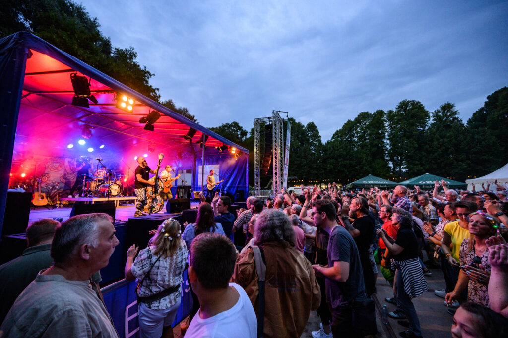 Vor einer Bühne im Schlosspark Wickrath feiert eine große Menschenmenge ein Konzert. Viele Hände sind in der Luft, während die Band unter farbigen Lichtern spielt. Im Hintergrund sind Zelte und Bäume zu sehen, die dem Fest am See in Mönchengladbach eine besondere Atmosphäre geben.