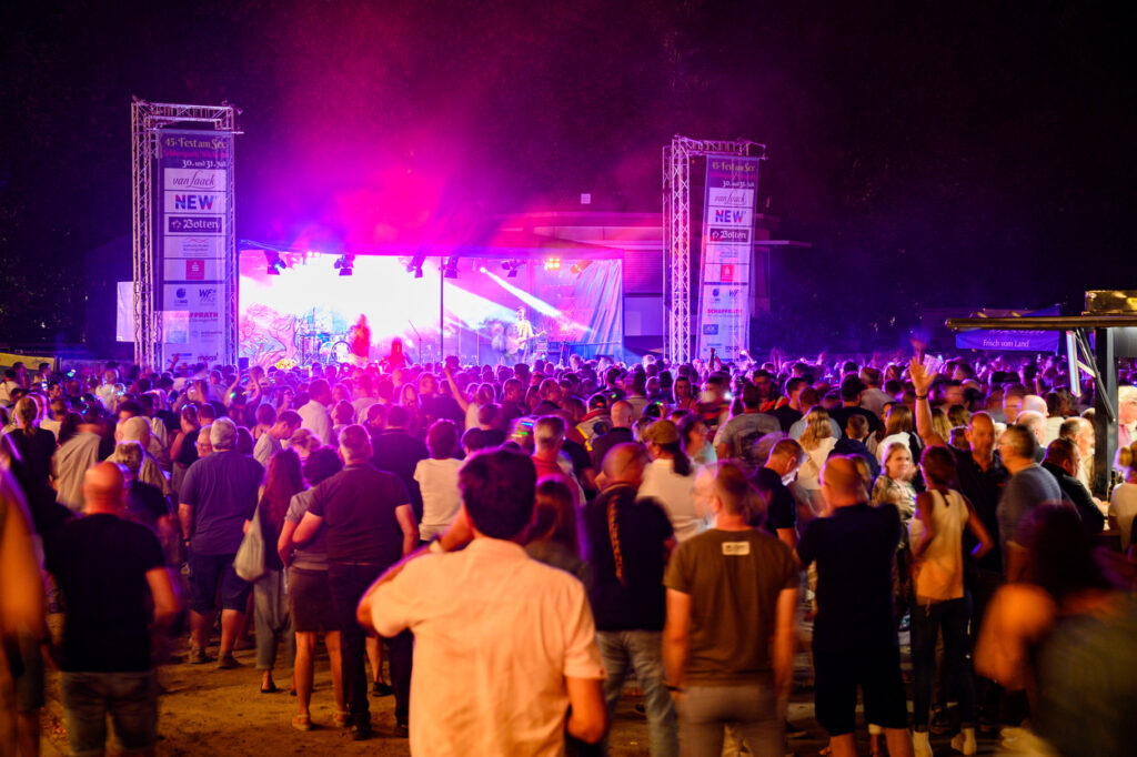 Eine große Menschenmenge versammelt sich vor einer Bühne im Schlosspark Wickrath bei Nacht. Die Bühne ist in pinkes Licht getaucht, während viele Gäste das Konzert verfolgen und sich durch das Gelände bewegen.
