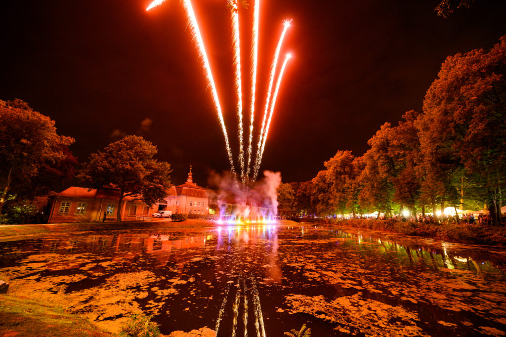 Ein Feuerwerk erleuchtet den Himmel über dem Schlosspark Wickrath und spiegelt sich im Wasser des Teichs. Im Hintergrund sind das Schloss und beleuchtete Bäume zu sehen, während das Festgelände stimmungsvoll erstrahlt.