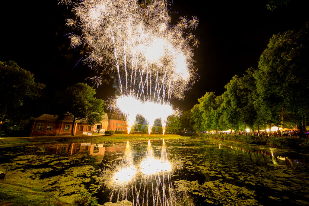 Ein großes Feuerwerk erleuchtet den Nachthimmel über dem Schlosspark Wickrath. Die goldenen Explosionen spiegeln sich im Wasser des Teichs, während Schloss, Bäume und das Festgelände stimmungsvoll beleuchtet sind.