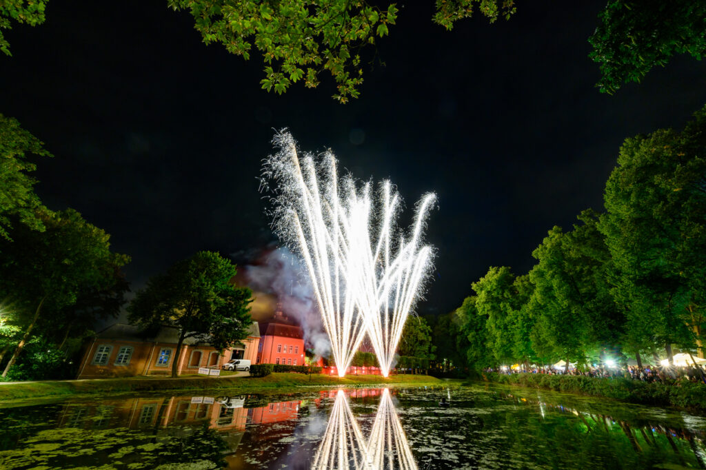 Mehrere helle Feuerwerksfontänen steigen gleichzeitig vor dem Schloss Wickrath in den Himmel. Die weißen Lichtstrahlen spiegeln sich klar im Wasser, umgeben von Bäumen und dem nächtlichen Park.