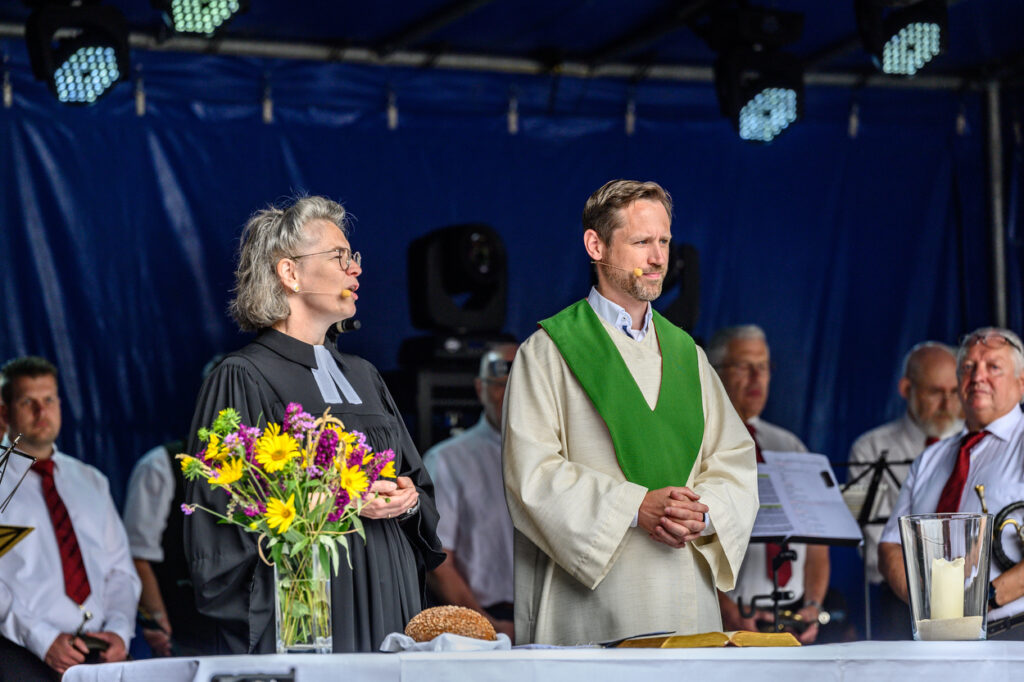 Eine Pfarrerin und ein Priester stehen auf einer Bühne bei einem Gottesdienst im Freien. Vor ihnen stehen Blumen, Brot und eine Kerze, im Hintergrund ist ein Chor zu sehen.