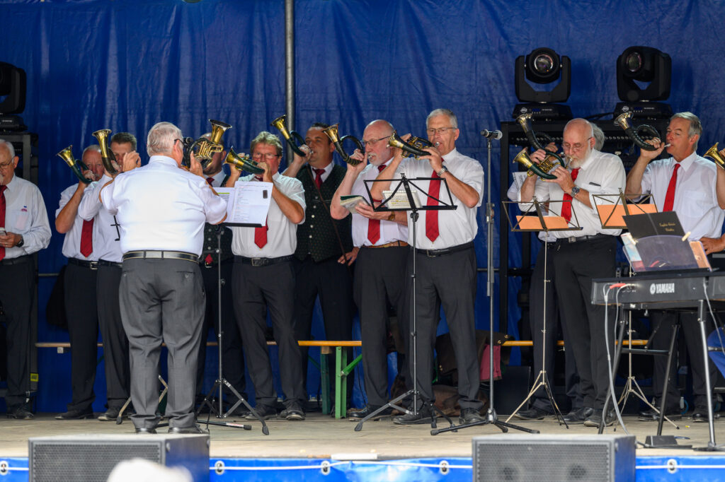 Eine Gruppe von Musikern in weißen Hemden und roten Krawatten spielt gemeinsam auf einer Bühne. Ein Dirigent steht mit dem Rücken zum Publikum vor ihnen.
