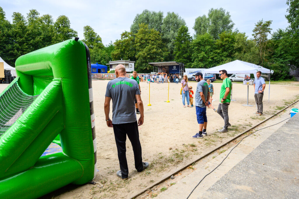 Sandfläche mit Eventbereich, Menschen stehen vor einer kleinen Bühne, im Vordergrund ein großes grünes aufblasbares Spielgerät.