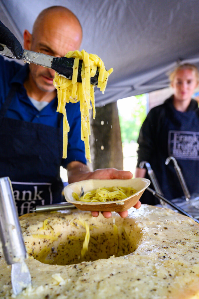 Frisch zubereitete Pasta wird aus einem großen Käselaib auf einen Teller gehoben, Koch im Vordergrund.