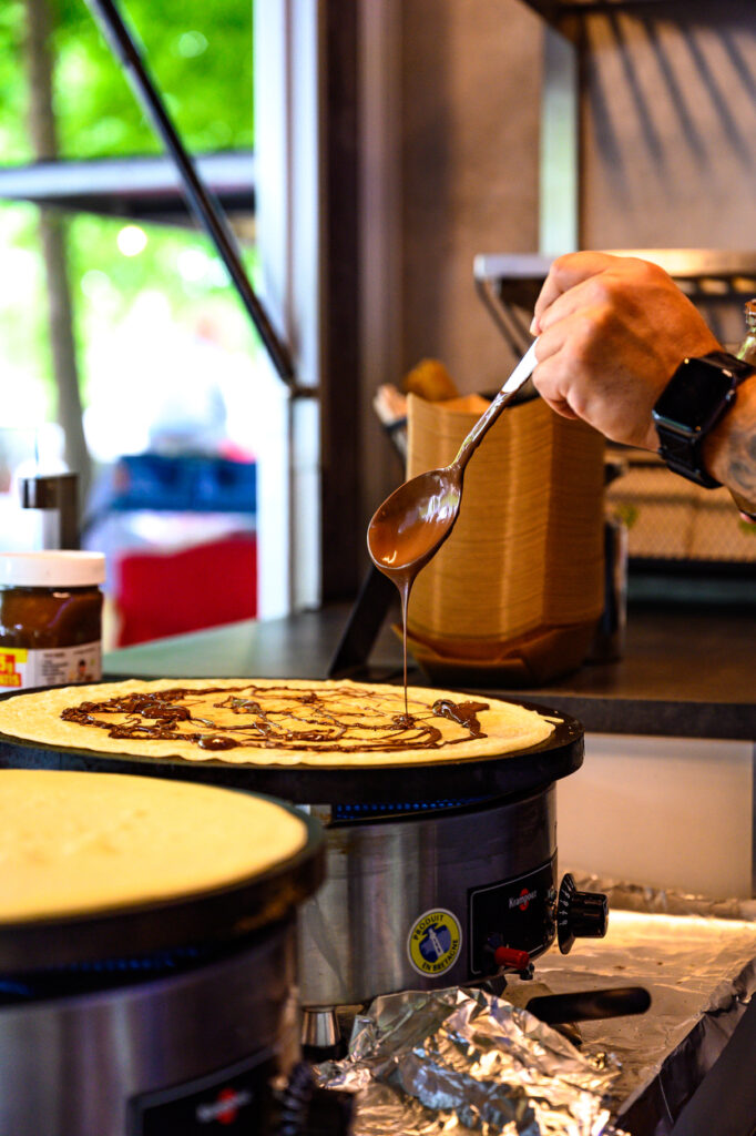 Crêpe wird auf einer heißen Platte mit Schokolade bestrichen, Hand mit Löffel im Vordergrund.
