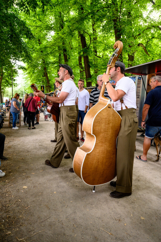 Musiker mit Kontrabass und Gitarre spielen auf einem Weg im Grünen, Publikum steht und schaut zu.