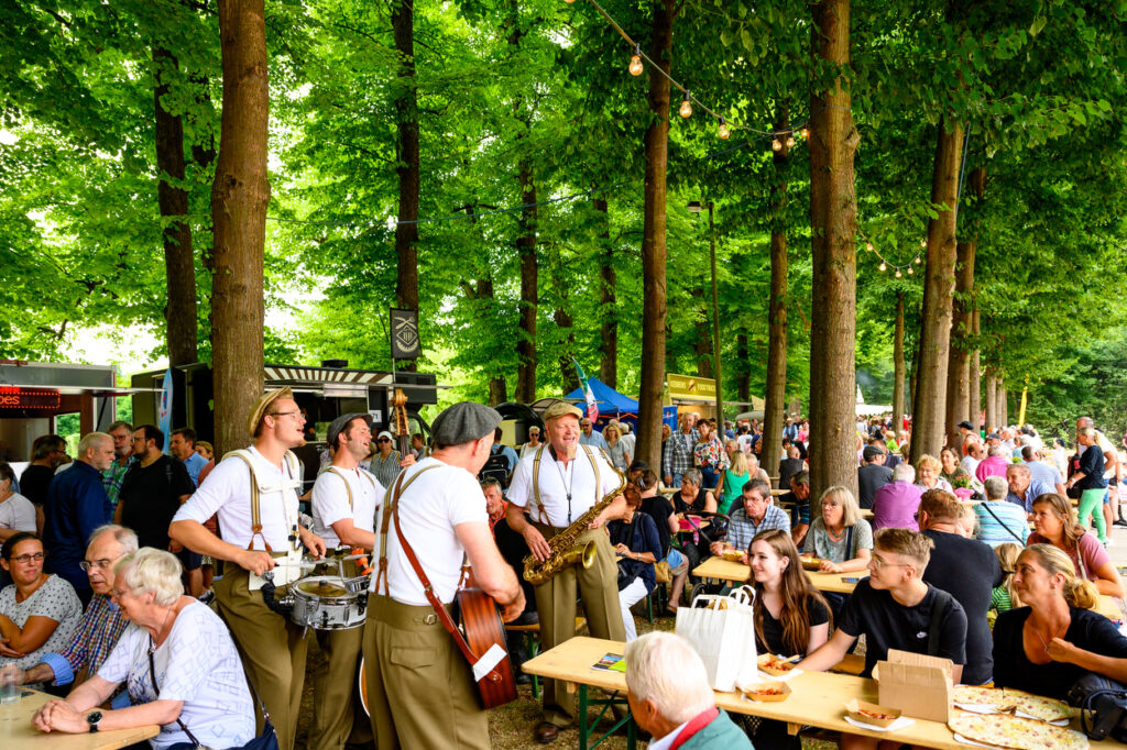 Menschen sitzen an langen Holztischen unter hohen Bäumen im Schlosspark Wickrath und essen gemeinsam. Eine kleine Band mit Saxophon, Kontrabass, Gitarre und Schlagzeug spielt zwischen den Tischen. Lichterketten hängen zwischen den Bäumen, viele Besucher schauen zu oder unterhalten sich.