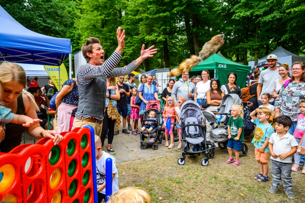Ein Entertainer wirft Gegenstände in die Luft, während eine Gruppe von Kindern und Erwachsenen zuschaut. Einige Kinder stehen vorne mit Kinderwagen und reagieren begeistert.