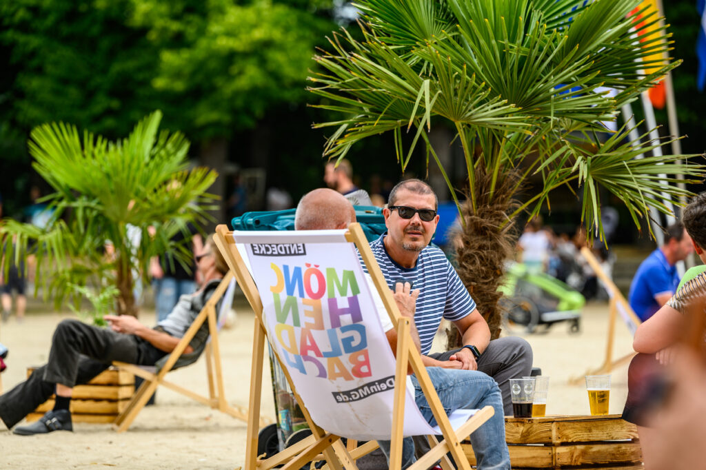 Ein Mann sitzt in einem Liegestuhl im Sand und schaut zur Kamera. Neben ihm stehen kleine Palmen und Getränke auf Holzkisten. Weitere Menschen entspannen im Hintergrund.