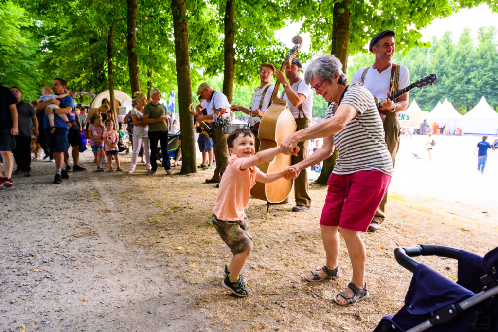 Ein Kind tanzt mit einer älteren Frau unter Bäumen im Park. Im Hintergrund spielt eine Band mit Kontrabass und Gitarre, während andere Besucher stehen und zuschauen.