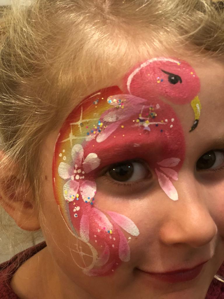 Ein Kind mit pinker Flamingo-Gesichtsbemalung und glitzernden Details lächelt.