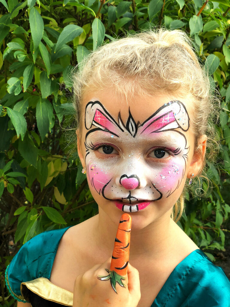 Ein Kind mit Hasen-Gesichtsbemalung hält einen bemalten Finger als Karotte vor das Gesicht.
